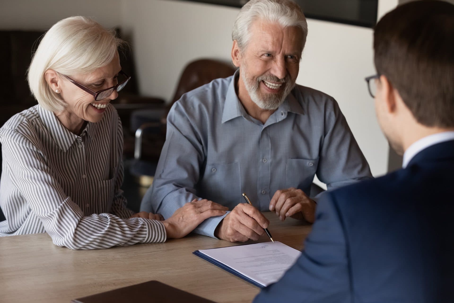 An older couple sit across a wooden conference table from an attorney in a navy suit, smiling as they sign estate planning documents together.