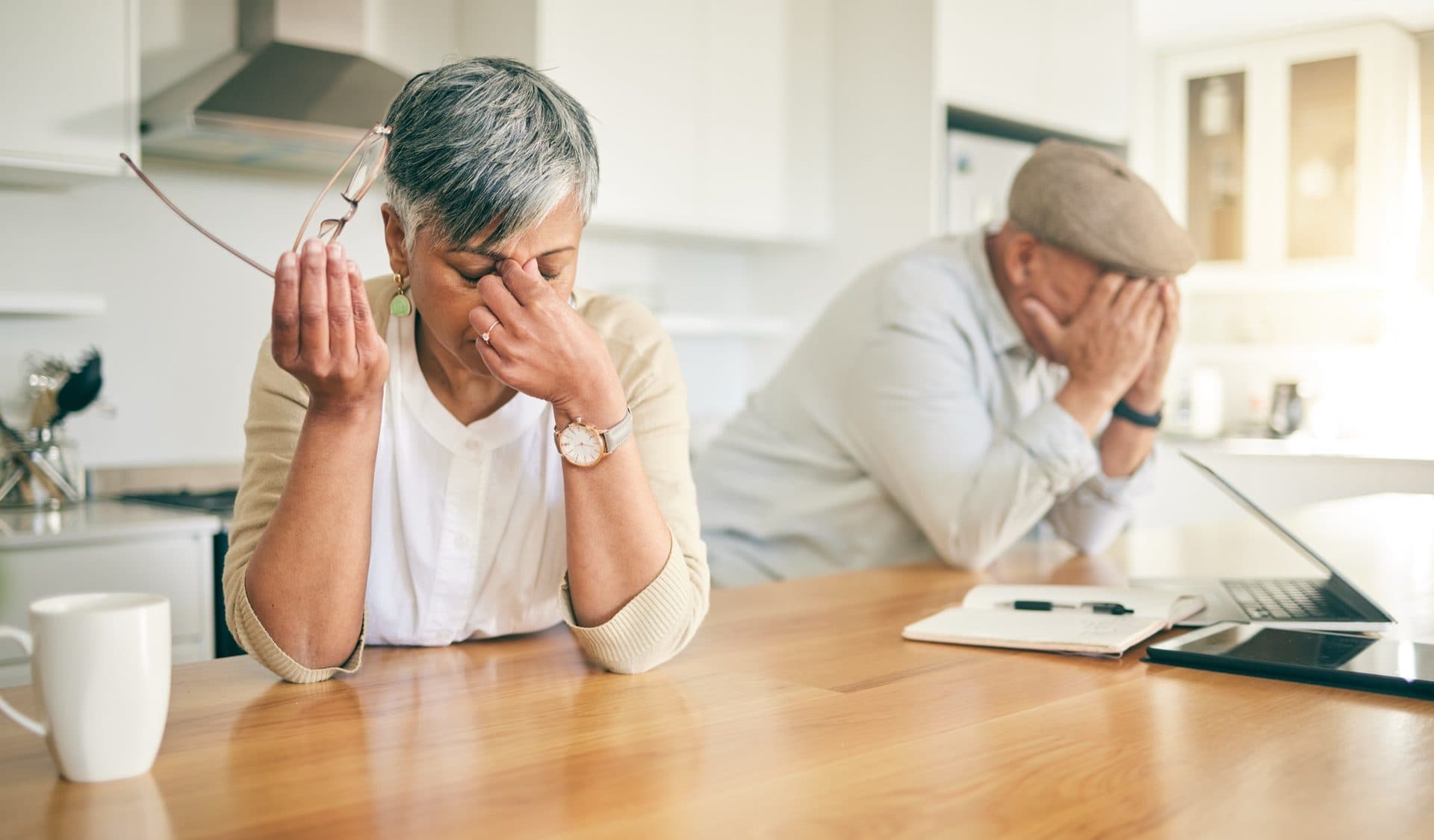 A grey-haired woman rubs her temple while sitting at a kitchen table; in the background a man holds his head in his hands. Both look stressed and overwhelmed.